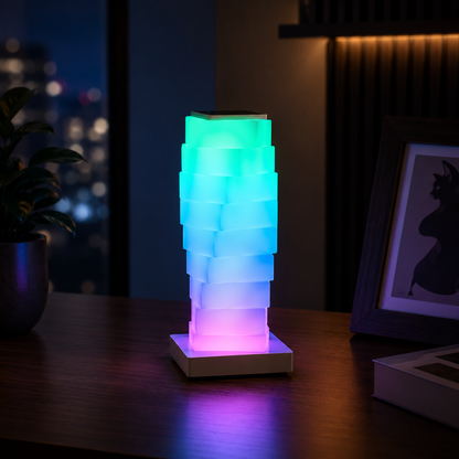 A modern LED block lamp with a tiered, stacked geometric design glows with a multicolor gradient transitioning from green to blue to pink, sitting on a dark wooden table in a dimly lit room.