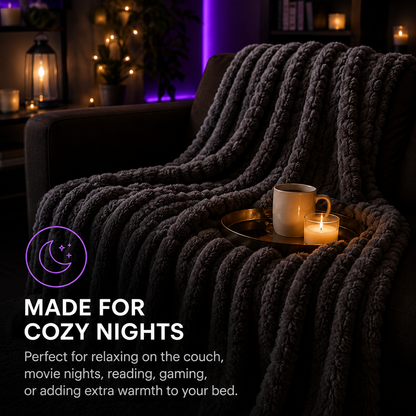 A thick, chunky knit throw blanket in dark charcoal gray draped over a couch in a cozy living room setting, with a mug and candle on a tray. Text on the image reads 'MADE FOR COZY NIGHTS' with descriptive copy below.